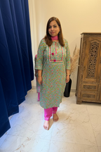A woman stands indoors wearing a light green printed kurta with pink and orange embroidery and bright pink pants, posed in front of a mirror in a bedroom-like space with a bed, curtains, and tiled floor.