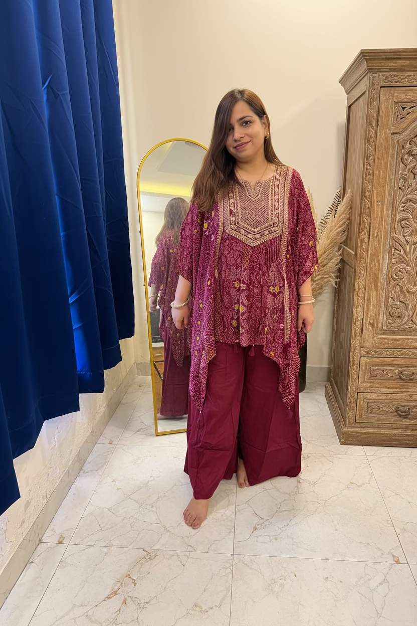 A woman stands barefoot in a bedroom-like interior wearing a deep magenta embroidered tunic with matching wide-leg pants. A full-length mirror, blue curtain, bed, and tiled floor are visible in the room.