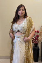 A woman modeling a white and gold traditional Indian outfit with a heavily embroidered cream and gold blouse, draped white skirt, and a sheer gold-patterned dupatta, standing in front of a mirror.