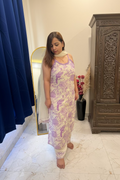 A woman stands barefoot in a bedroom wearing a light cream or off-white sleeveless kurta with a purple bird pattern and a matching light dupatta draped over her shoulder. The room has a cream wall, a blue curtain on the left, a mirror, and a bed with floral bedding on the right.