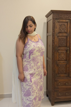 A woman wearing a sleeveless cream-colored dress with a purple peacock and floral toile pattern, styled with a white dupatta, standing in an indoor setting.