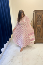 A woman wearing a pink and white patterned kaftan dress with a matching belt, standing in a bedroom setting next to a bed with a floral-patterned sheet.