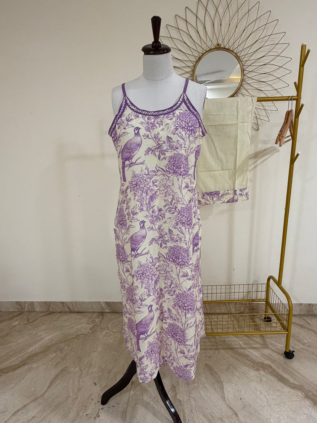 A sleeveless midi dress with a purple toile-style print on a cream background, displayed on a mannequin. A matching cream towel with a purple border is draped over a gold clothing rack in the background.