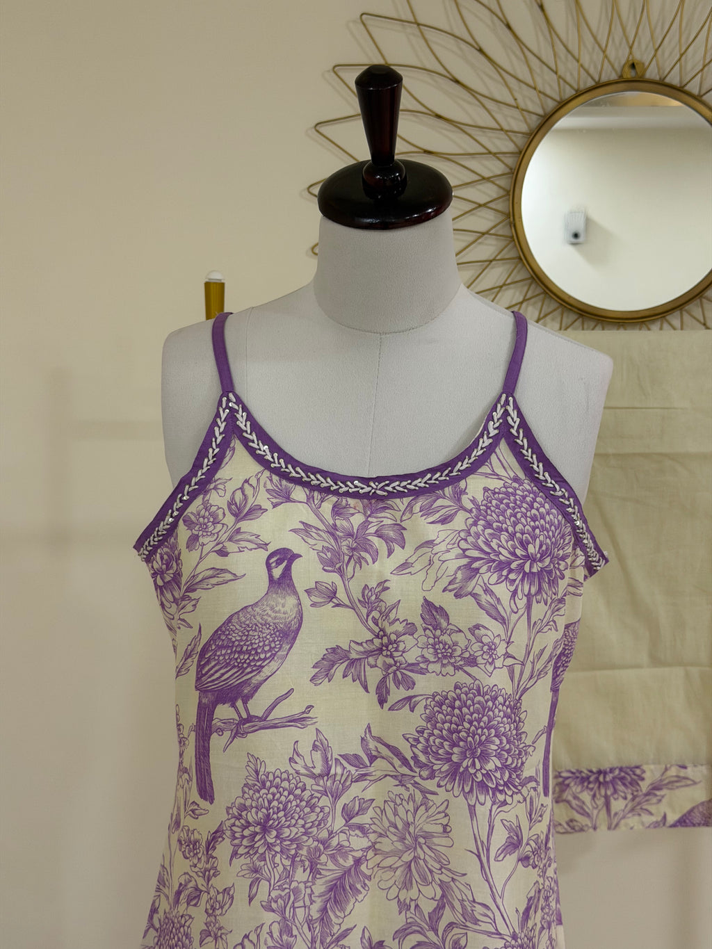 A sleeveless camisole tank top displayed on a dress form mannequin. The top features a purple and cream toile-style print with a bird and floral motifs, and a purple braided trim along the neckline.