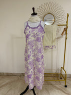 A sleeveless midi dress with a purple and white toile-style print featuring birds and floral motifs, displayed on a mannequin. A cream scarf is wrapped around the neck, and a gold clothing rack with a sunburst mirror and folded cream fabric stands behind it against a plain wall.