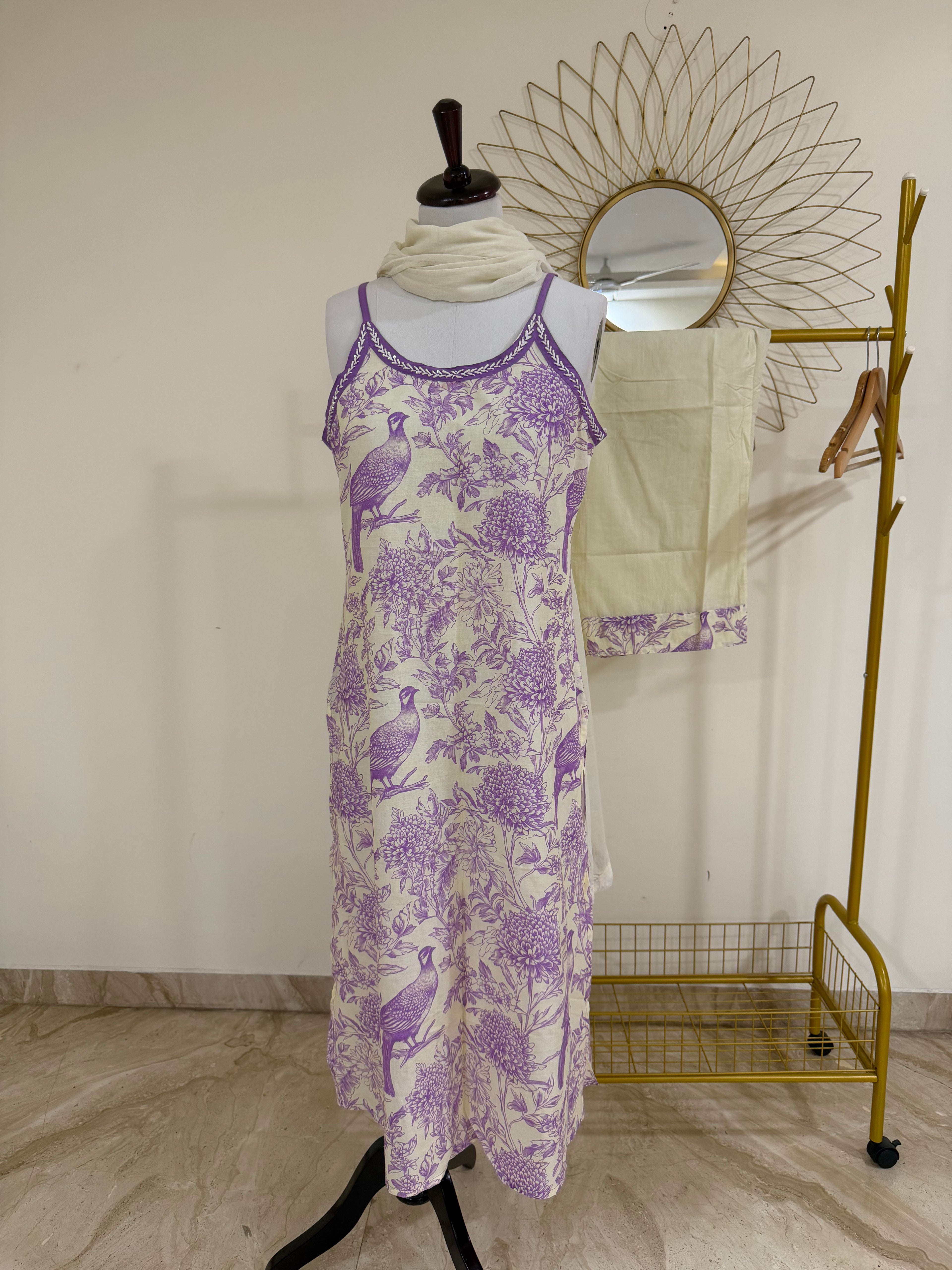 A sleeveless midi dress with a purple and white toile-style print featuring birds and floral motifs, displayed on a mannequin. A cream scarf is wrapped around the neck, and a gold clothing rack with a sunburst mirror and folded cream fabric stands behind it against a plain wall.