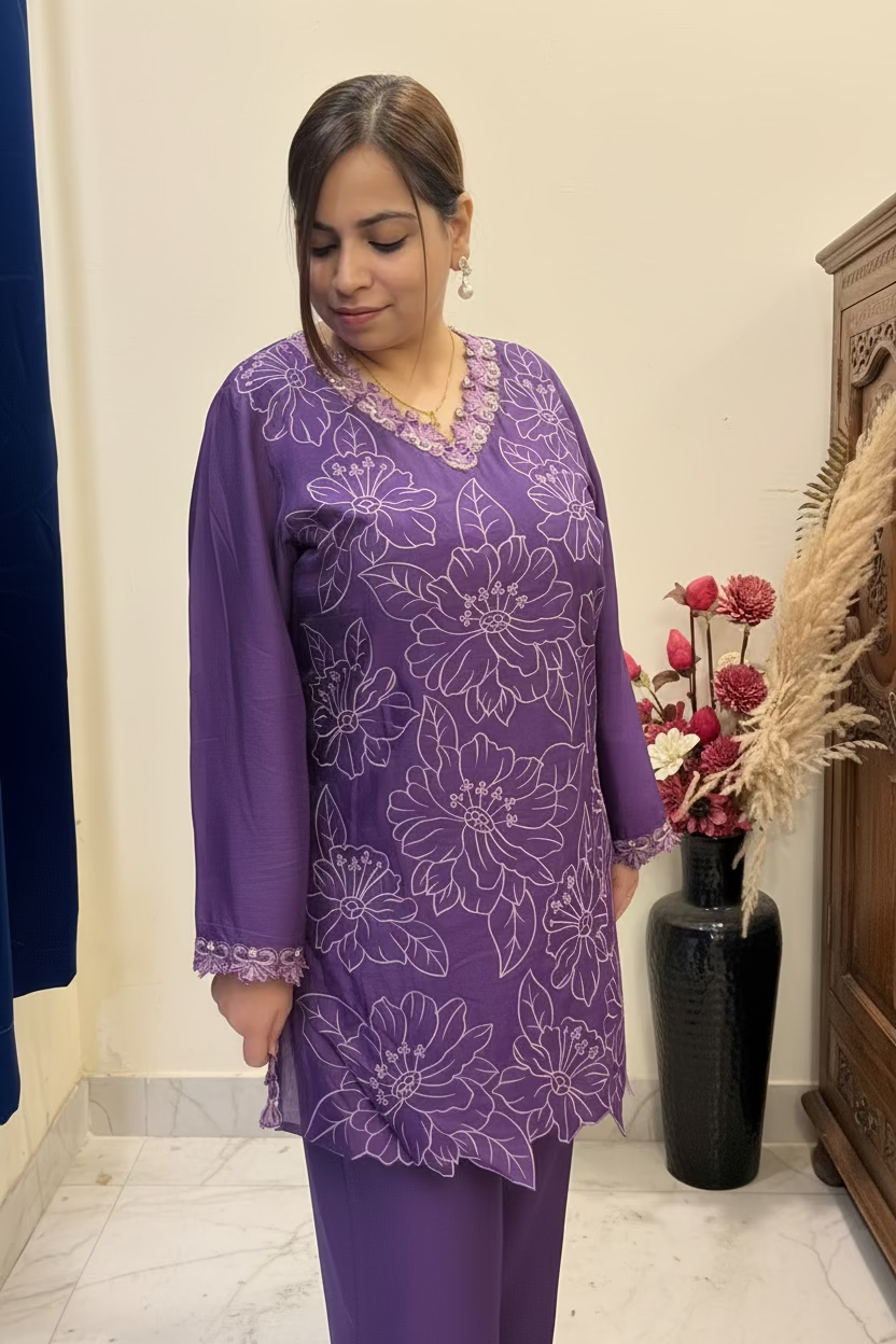 A woman wearing a purple embroidered kurta with matching pants, standing indoors in front of a mirror. The kurta features light purple floral embroidery and scalloped edges.