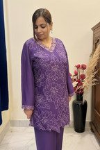 A woman wearing a purple embroidered kurta with matching pants, standing indoors in front of a mirror. The kurta features light purple floral embroidery and scalloped edges.