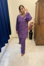 A woman wearing a purple embroidered tunic and matching pants stands barefoot in front of a mirror in a bedroom setting.