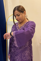 A woman wearing a purple embroidered kurta with floral patterns and bell sleeves, looking down at her wrist while wearing a black watch.