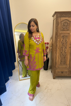A woman modeling a bright lime green traditional Indian outfit with pink floral embroidery and silver mirror work on the neckline, standing in a bedroom setting with a mirror, bed, and blue curtain visible.
