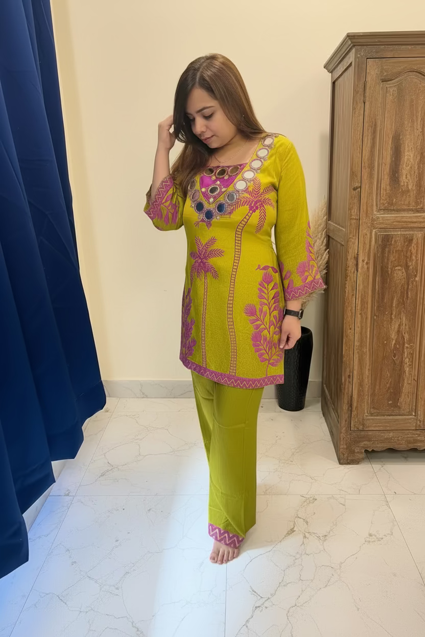 A woman modeling a bright yellow-green kurta and trouser set with pink floral embroidery and colorful beaded neckline details, standing in front of a mirror in an indoor room with a wooden cabinet and blue curtain.