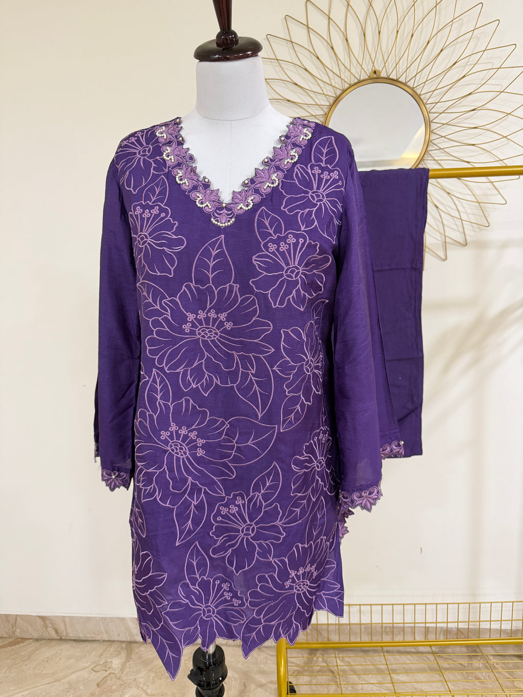 A purple embroidered tunic dress with a matching draped fabric piece, displayed on a mannequin in an indoor setting with a decorative gold sunburst mirror in the background.