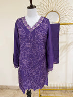 A purple embroidered tunic dress with a matching draped fabric piece, displayed on a mannequin in an indoor setting with a decorative gold sunburst mirror in the background.