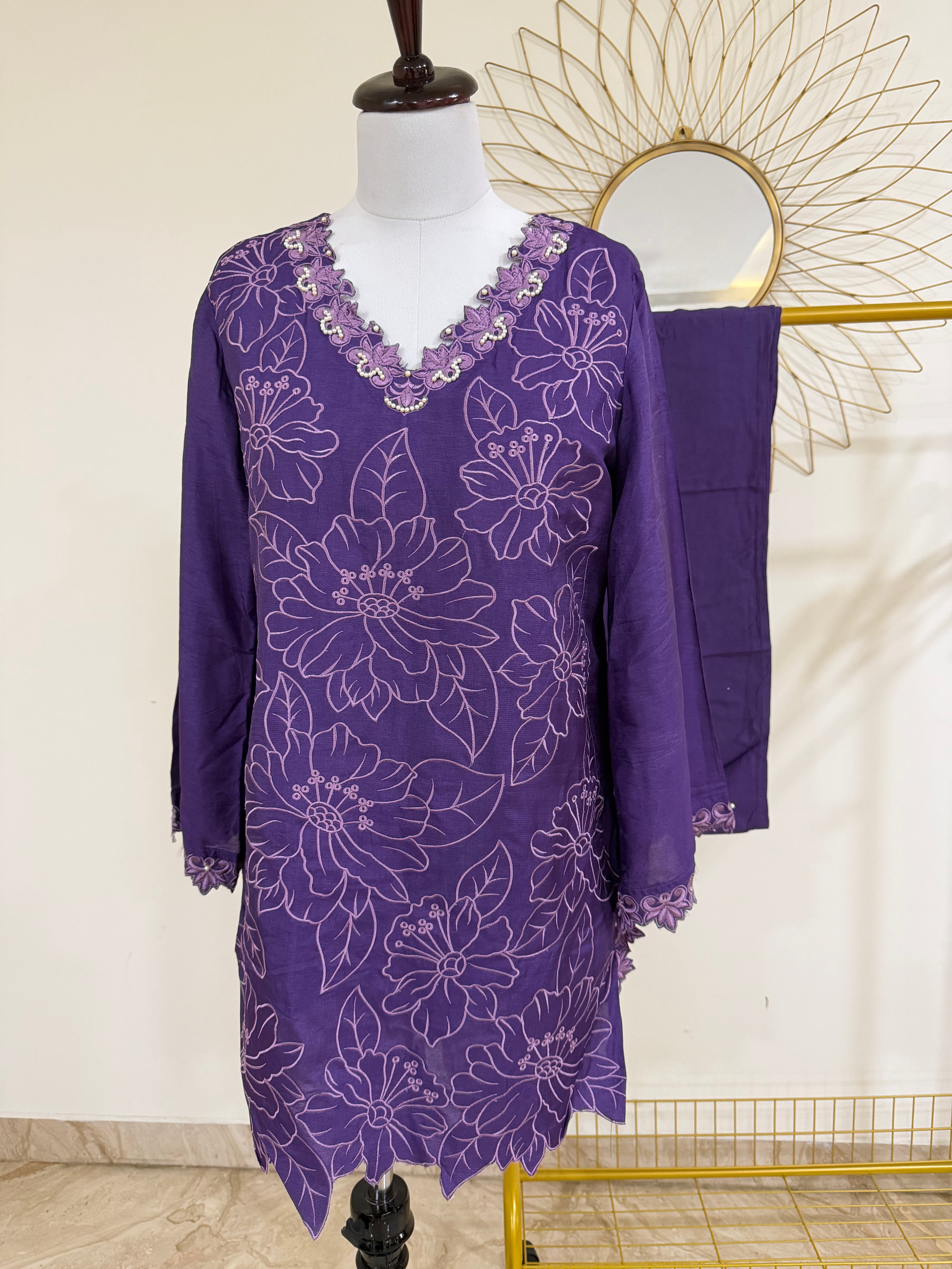 A purple embroidered tunic dress with a matching draped fabric piece, displayed on a mannequin in an indoor setting with a decorative gold sunburst mirror in the background.