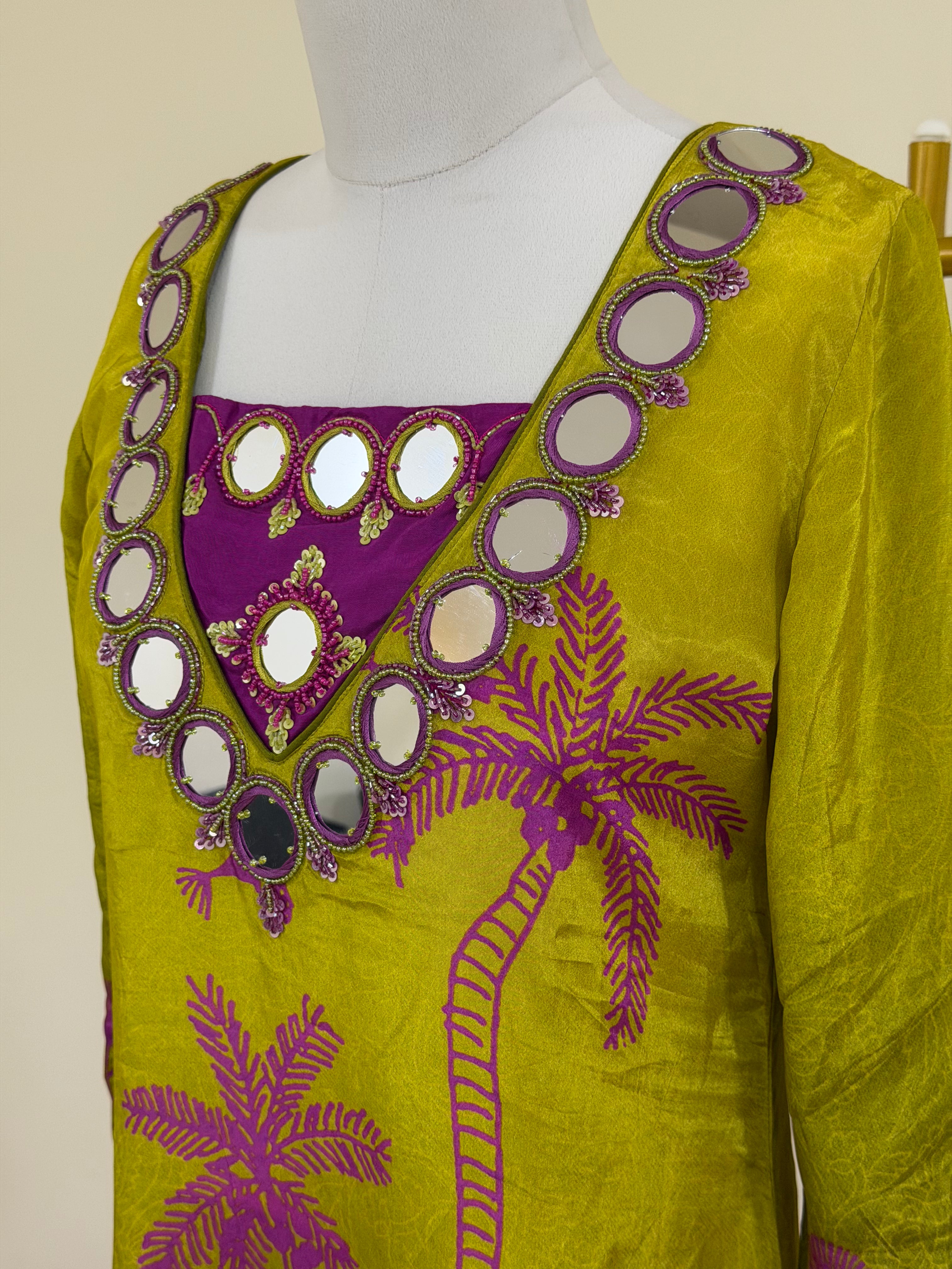 A close-up of a yellow-green embroidered kurta on a white mannequin torso. The garment features a purple inner neckline with circular mirror work, a purple chest panel with gold mirror accents, and purple palm tree embroidery on the front.