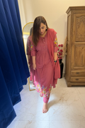 A woman wearing a pink traditional Indian salwar kameez set with floral embroidery, standing in a bedroom setting with a bed, curtains, and a mirror visible.