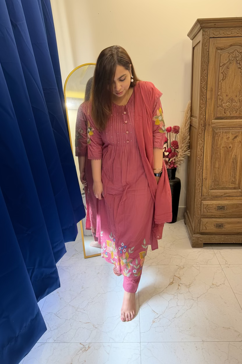 A woman wearing a pink traditional Indian salwar kameez set with floral embroidery, standing in a bedroom setting with a bed, curtains, and a mirror visible.