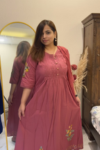 A woman wearing a dusty rose pink embroidered kurta dress with three-quarter sleeves and a buttoned front, standing in front of a mirror in an indoor setting.