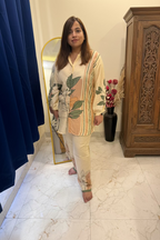 A woman modeling a cream-colored two-piece pajama set with green leaf prints and vertical stripes on the tunic, standing in a bedroom setting.