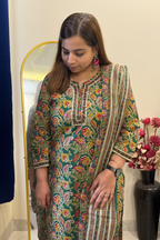 A woman wearing a green printed kurta with colorful floral patterns and a matching striped dupatta, standing indoors next to a mirror and a vase of flowers.