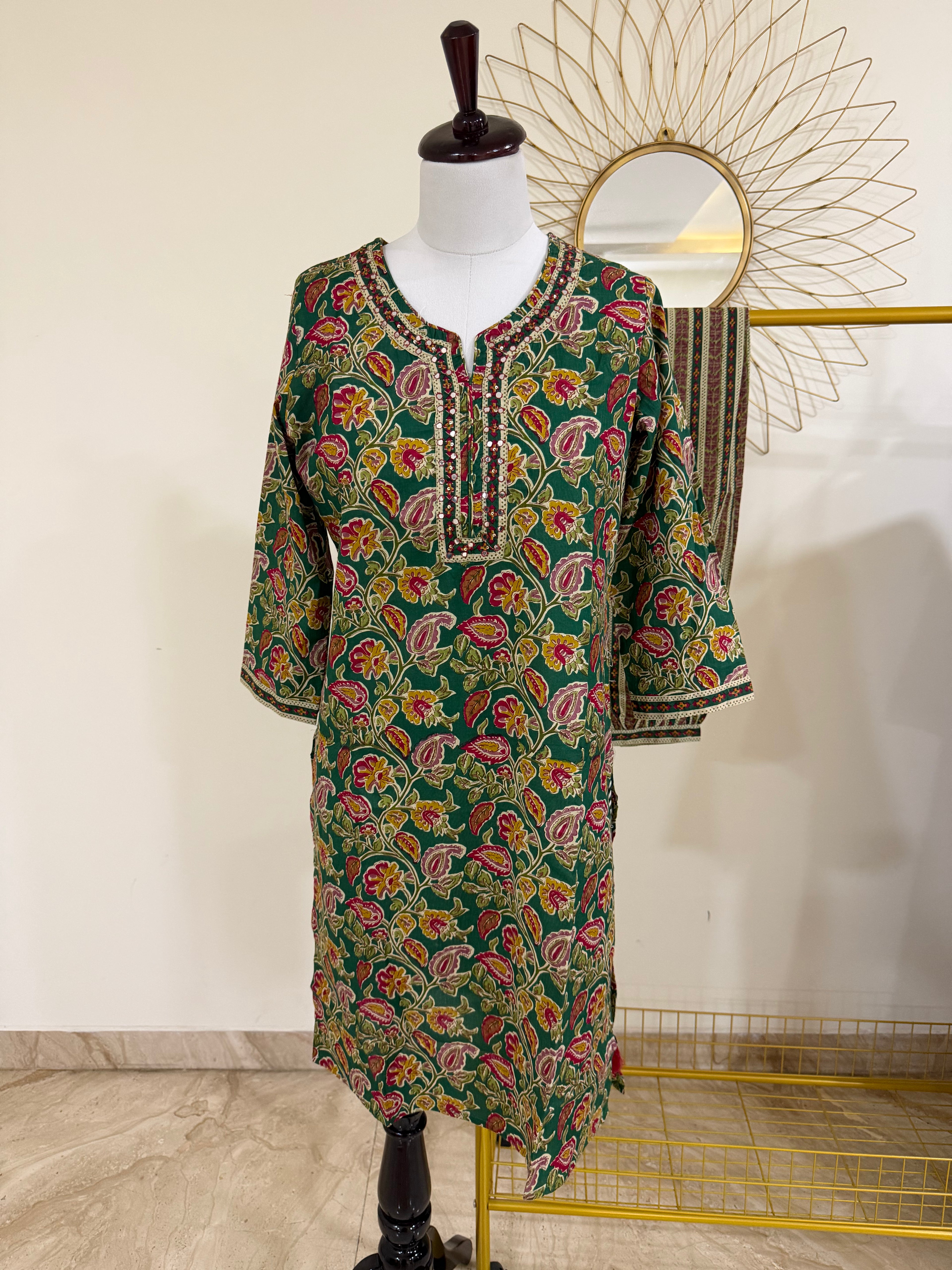 A green printed tunic dress with a colorful floral and paisley pattern, displayed on a mannequin. The dress features a V-neckline with embroidered detailing in black, gold, and red.