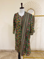 A three-piece women's ethnic suit set displayed on a mannequin, featuring a green printed kurta with colorful paisley motifs, matching trousers, and a striped dupatta draped over the shoulder.