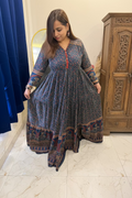 A woman wearing a long-sleeved, tiered maxi dress with a blue and red floral print and a patterned border hem, standing in a bedroom setting.