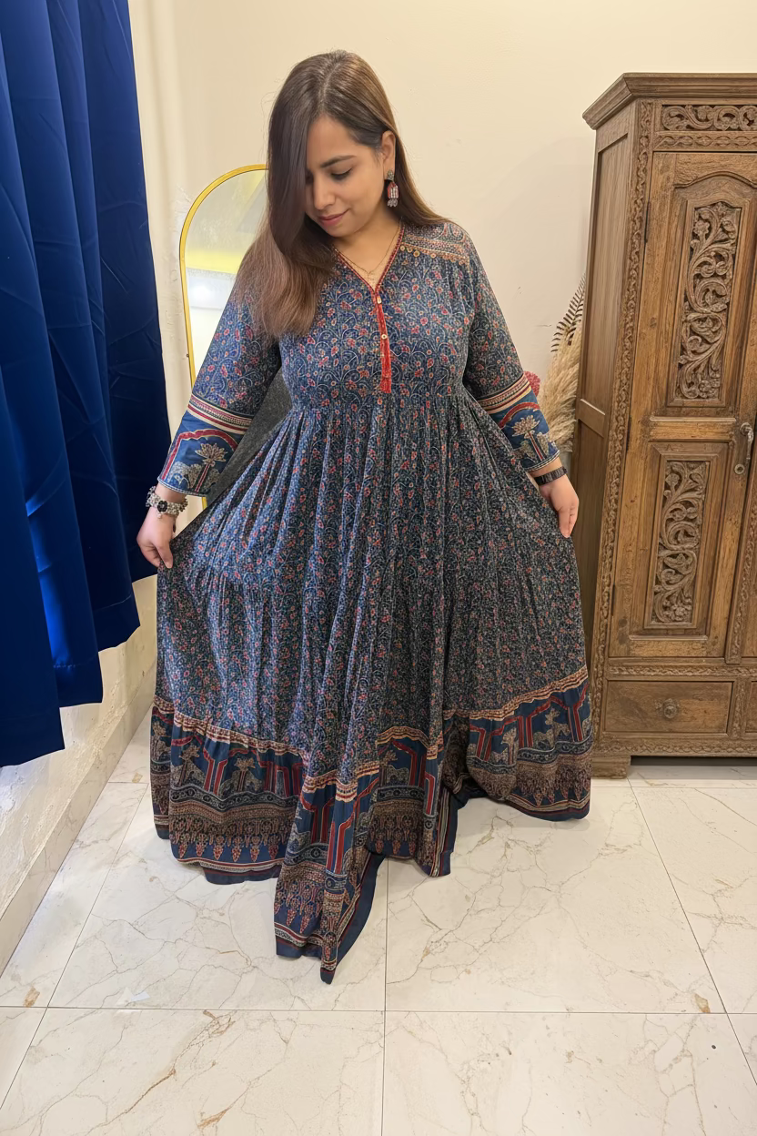 A woman wearing a long-sleeved, tiered maxi dress with a blue and red floral print and a patterned border hem, standing in a bedroom setting.