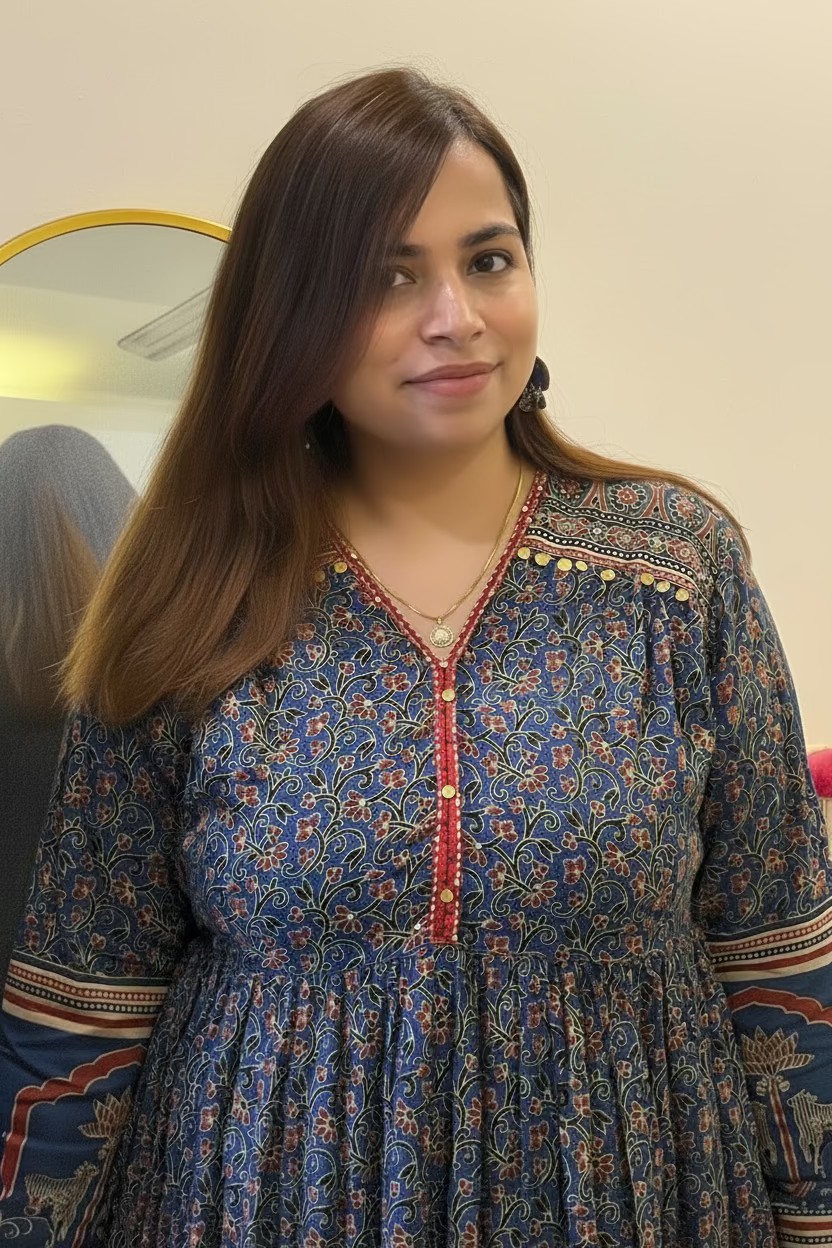 A woman wearing a blue floral printed kurta with red and gold accents, standing indoors in front of a cream-colored wall.