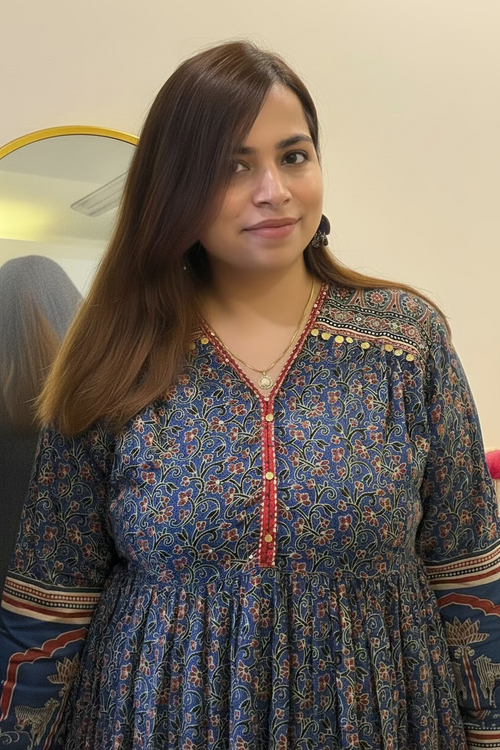 A woman wearing a blue floral printed kurta with red and gold accents, standing indoors in front of a cream-colored wall.
