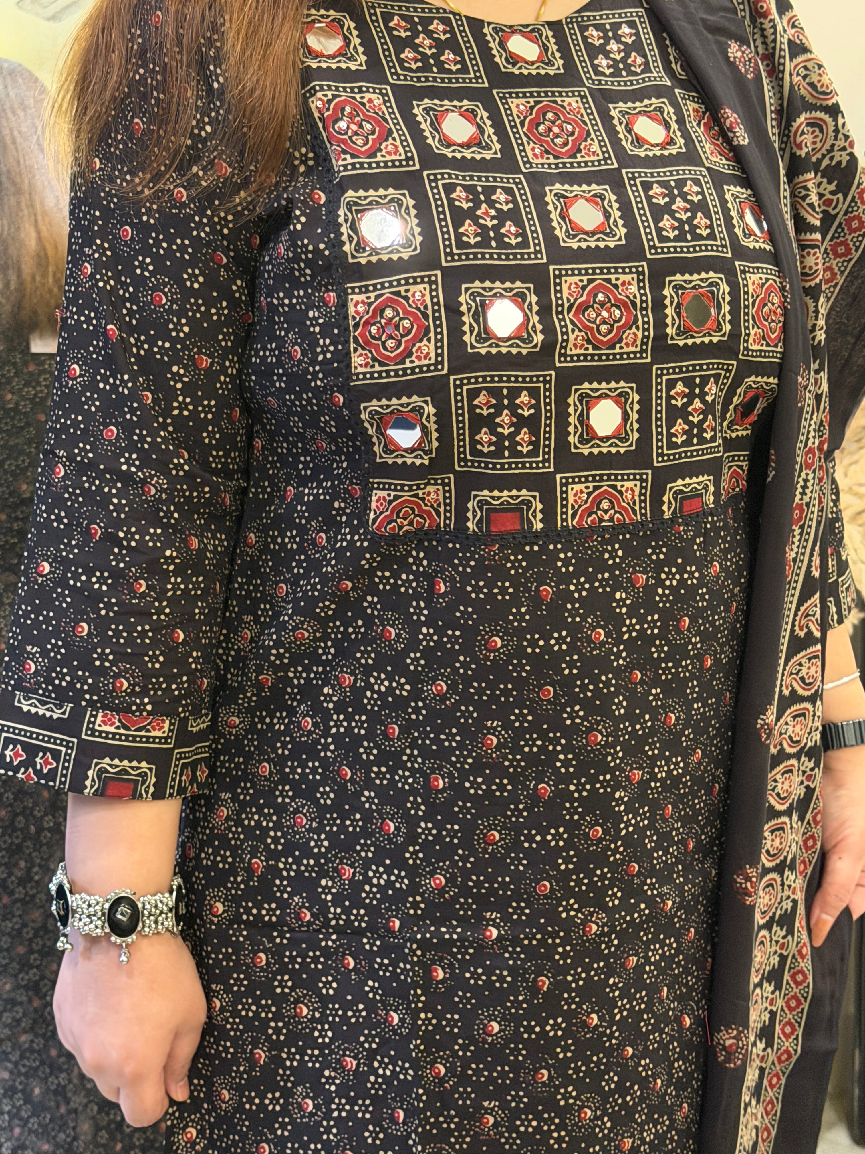 A close-up of a woman wearing a black three-piece salwar kameez suit set with a printed pattern, featuring a printed kameez shirt with mirror work, matching trousers, and a dupatta.
