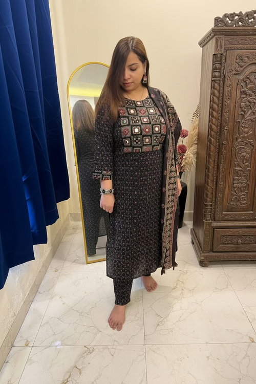 A woman modeling a dark brown printed salwar kameez set with a matching dupatta in a bedroom setting.