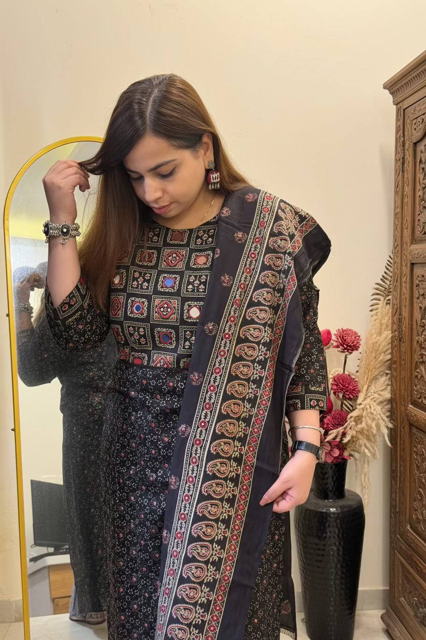 A woman wearing a dark charcoal gray printed salwar kameez suit with colorful embroidered borders and a matching dupatta, standing in front of a mirror in a home setting.