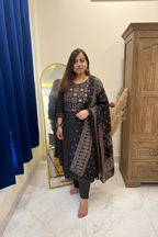A woman modeling a black Indian salwar kameez suit with intricate multicolor geometric patterns and a matching printed dupatta, standing in a bedroom setting.