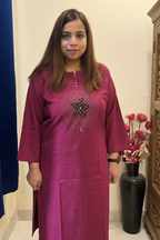 A woman wearing a magenta-pink kurta with a circular silver beaded embellishment on the chest, standing in an indoor setting with a beige wall, blue curtain, and a vase of flowers.