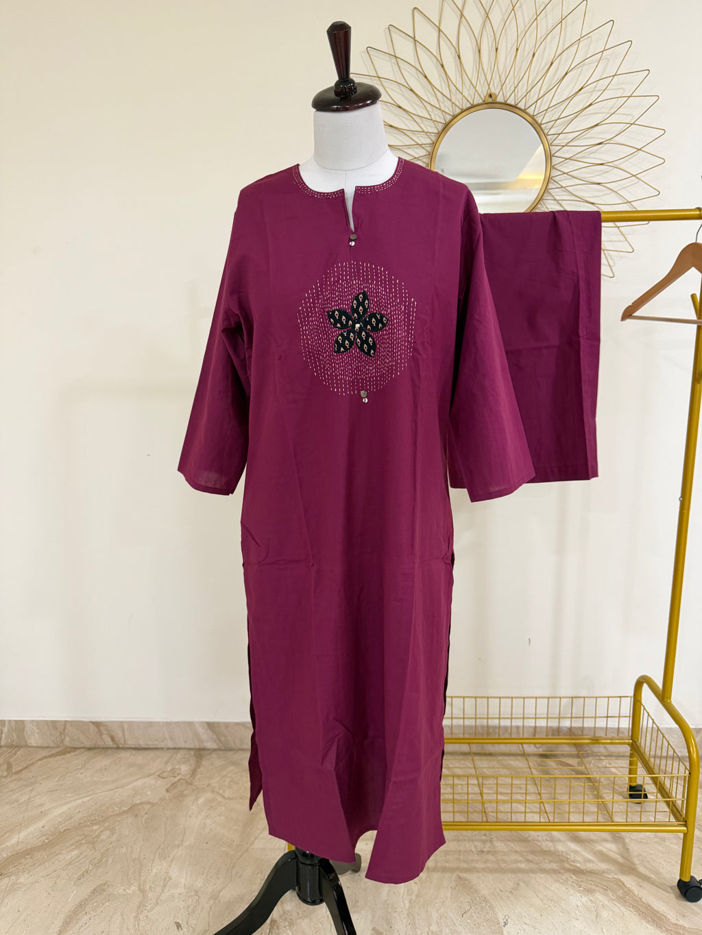 A magenta-purple long kurta displayed on a mannequin, featuring a circular embroidered star design on the chest. A matching pair of trousers is draped over a gold clothing rack in the background.