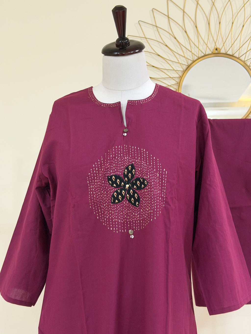 A magenta purple long-sleeve tunic top displayed on a white mannequin torso. The tunic features a round neckline with a split V-stitch, a central circular embellishment with a black and green floral motif, and small silver bead accents.