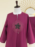 A magenta purple long-sleeve tunic top displayed on a white mannequin torso. The tunic features a round neckline with a split V-stitch, a central circular embellishment with a black and green floral motif, and small silver bead accents.