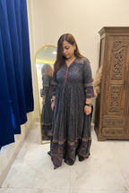 A woman wearing a long-sleeved, floor-length printed dress with a red embroidered neckline and multicolor patterned borders, standing in a bedroom setting.