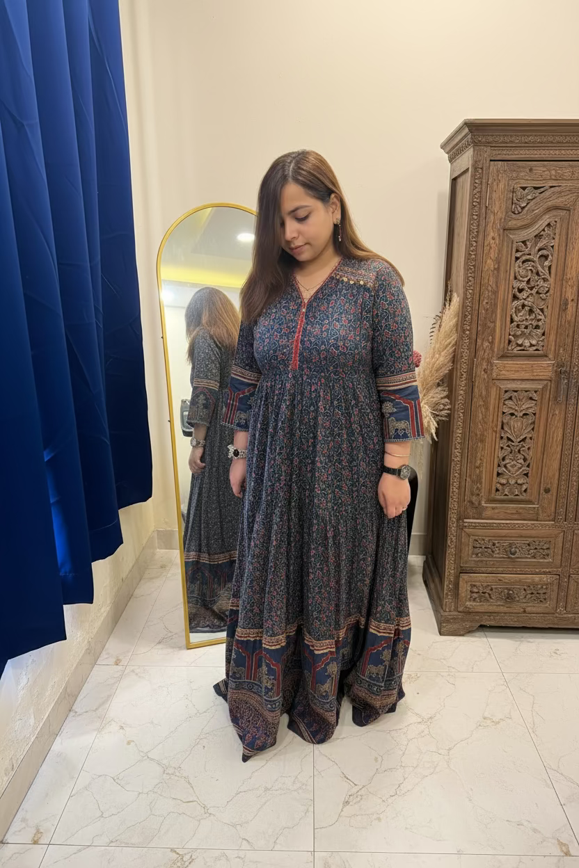 A woman wearing a long-sleeved, floor-length printed dress with a red embroidered neckline and multicolor patterned borders, standing in a bedroom setting.
