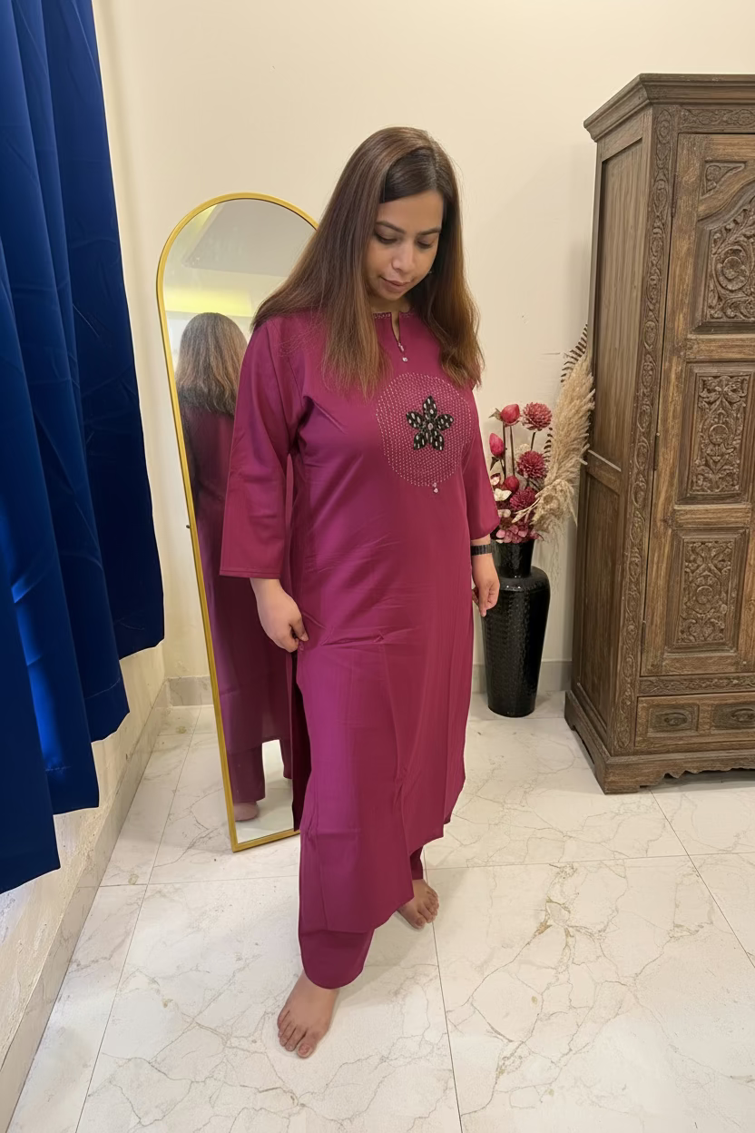 A woman wearing a magenta-purple kurta with a black embroidered chest motif, standing barefoot in a room with a marble floor, a wooden cabinet, and a blue curtain.